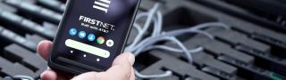Hand holds a phone with the FirstNet logo over a case of additional cell phones.