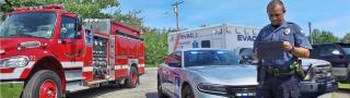 A police officer using a tablet stands in front of a fire truck, police car and ambulance.