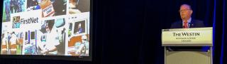 FirstNet Authority Board Chair Edward Horowitz stands behind a podium with a large screen featuring the FirstNet logo and images of first responders to his right.