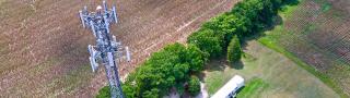 Aerial view of the top of a FirstNet cell site tower with a field and grove of trees in the background.