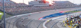 Race cars driving on the Iowa Speedway’s seven lane, oval racetrack in Newton, Iowa with thousands of spectators sitting in the surrounding stadium.