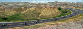 A scenic view of a winding road cutting through rocky hills in South Dakota, with a group of motorcyclists riding one behind the other along the curve of the road, surrounded by hills and rural terrain under a partly cloudy sky.