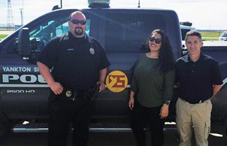 Two men and one woman stand in front of a Yankton Police truck. One of the men wears a police uniform.