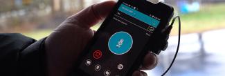 Hand holds FirstNet smartphone device with push-to-talk app displayed.