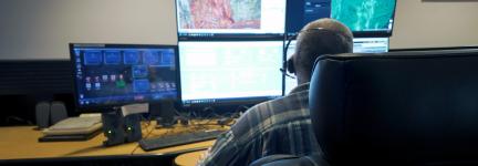 911 call-taker wearing a headset sitting at a desk in a communications center; multiple computer monitors showing maps, data, and emergency calls
